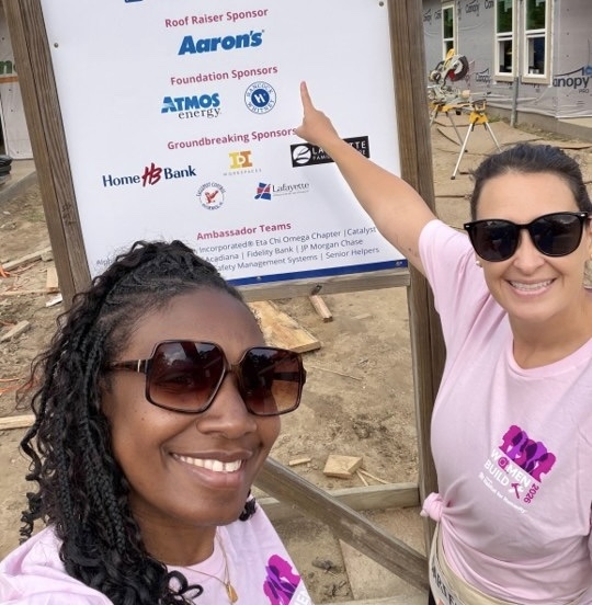 Two women wearing pink tees and sunglasses smile and point toward a large sign that reads ‘Habitat for Humanity – Women’s Build – Future Home Site’ followed by a list of ‘presented by’ companies, including Aaron’s as the ‘Raise the Roof Sponsor.’