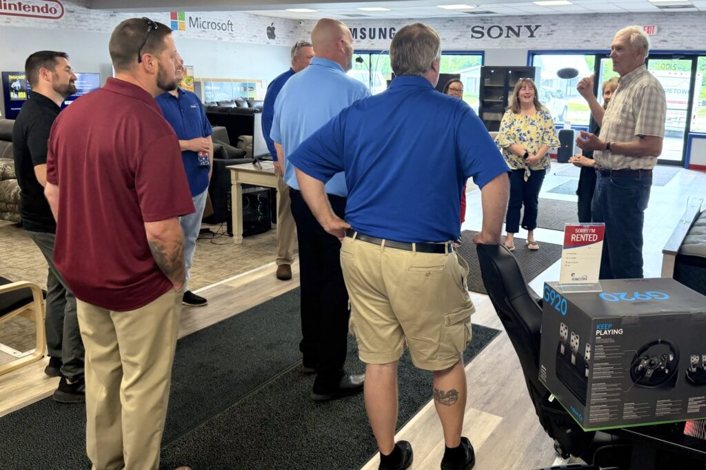 A group of men and women stand around a storefront showroom listening to an older man wearing a plaid shirt speak.