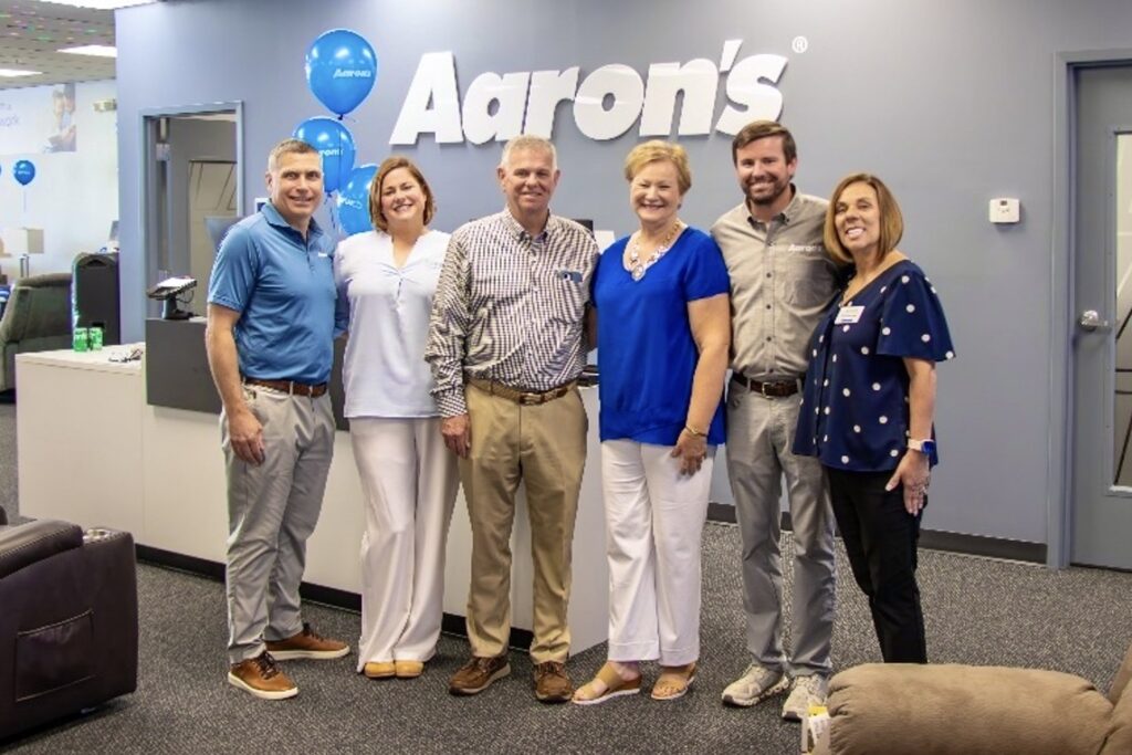 Several men and women stand together smiling in a showroom with a large Aaron’s sign and blue balloons behind them.