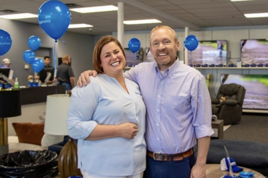 A woman and man stand together smiling on a store floor with blue balloons around them.