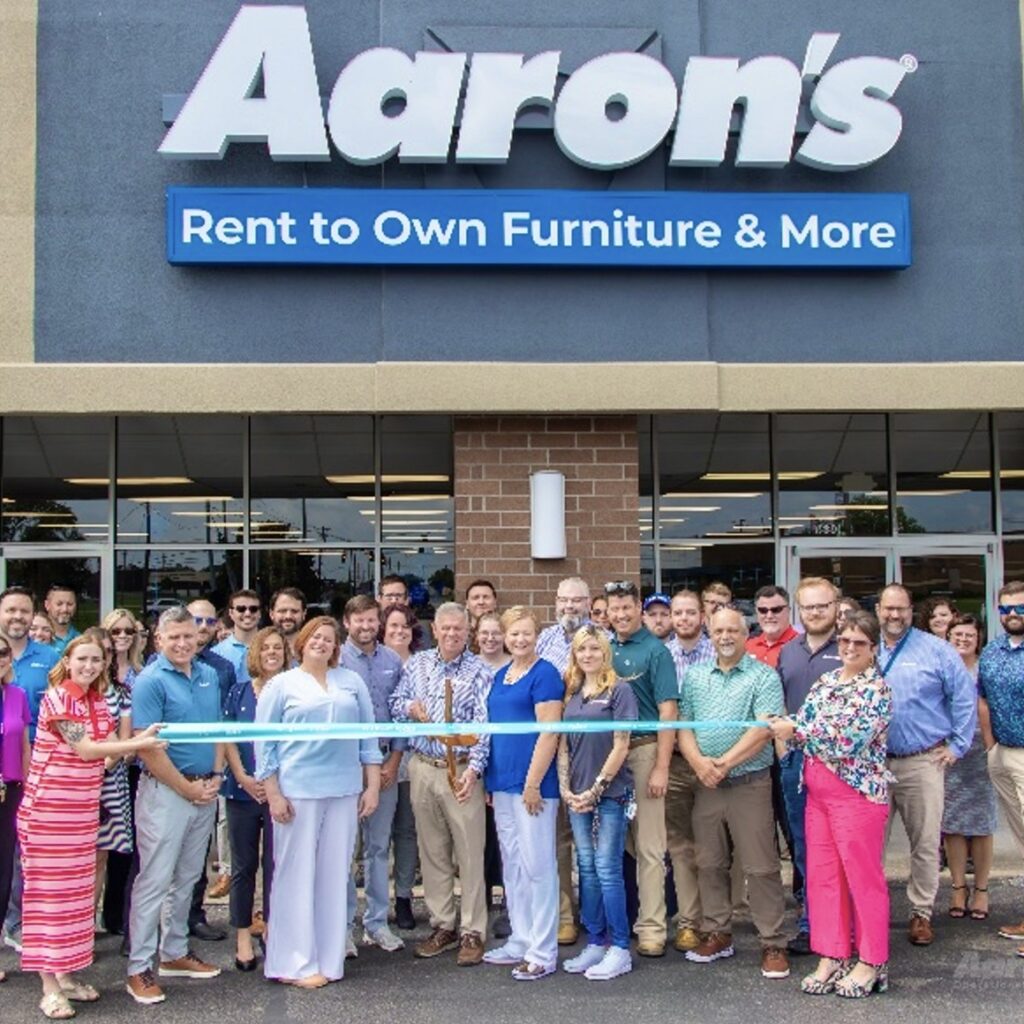 A large group of men and women stand smiling in front of a building with an Aaron’s sign above them. Two women hold a long, turquoise-colored ribbon in front of the group, while a man wearing a blue-patterned shirt and holding giant scissors prepares to cut the ribbon.