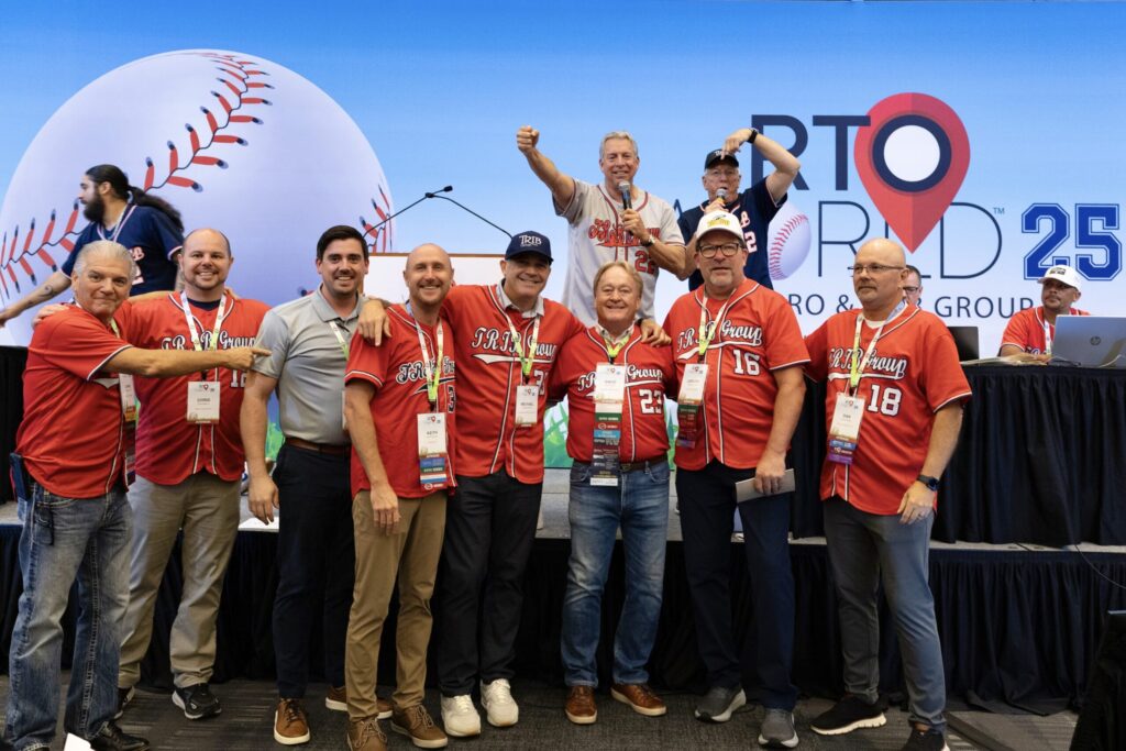 A group of men, most wearing red baseball jerseys reading “TRIB Group,” stand together smiling, with a stage behind them and a backdrop reading “RTO World 25.”