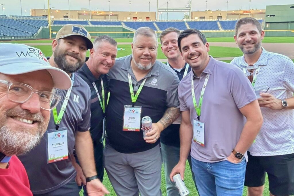 A group of men stand together smiling on the grassy diamond of a baseball park.