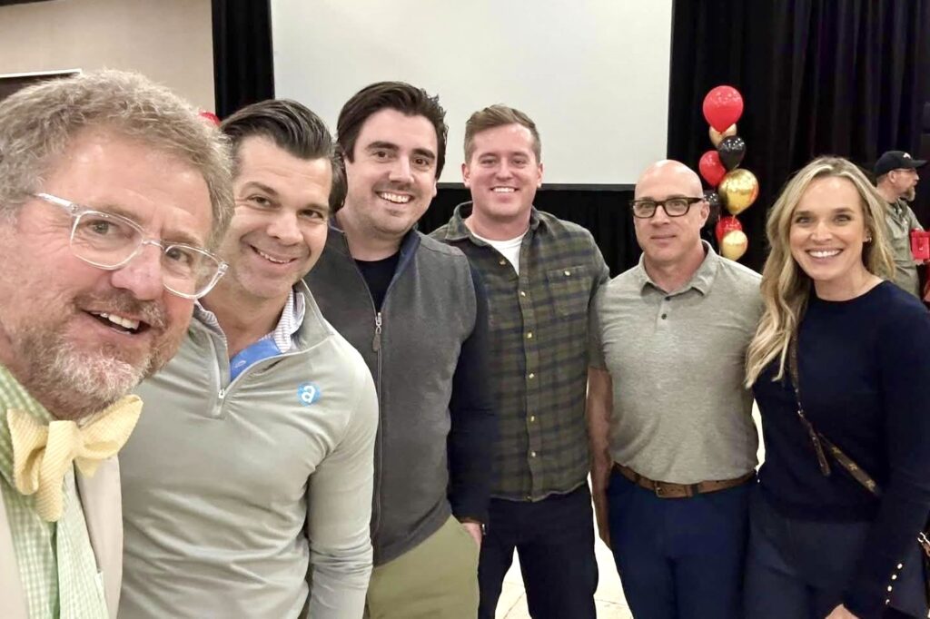 Four men and a blonde woman stand together smiling at an industry event, with bouquets of black, red, and gold balloons behind them.