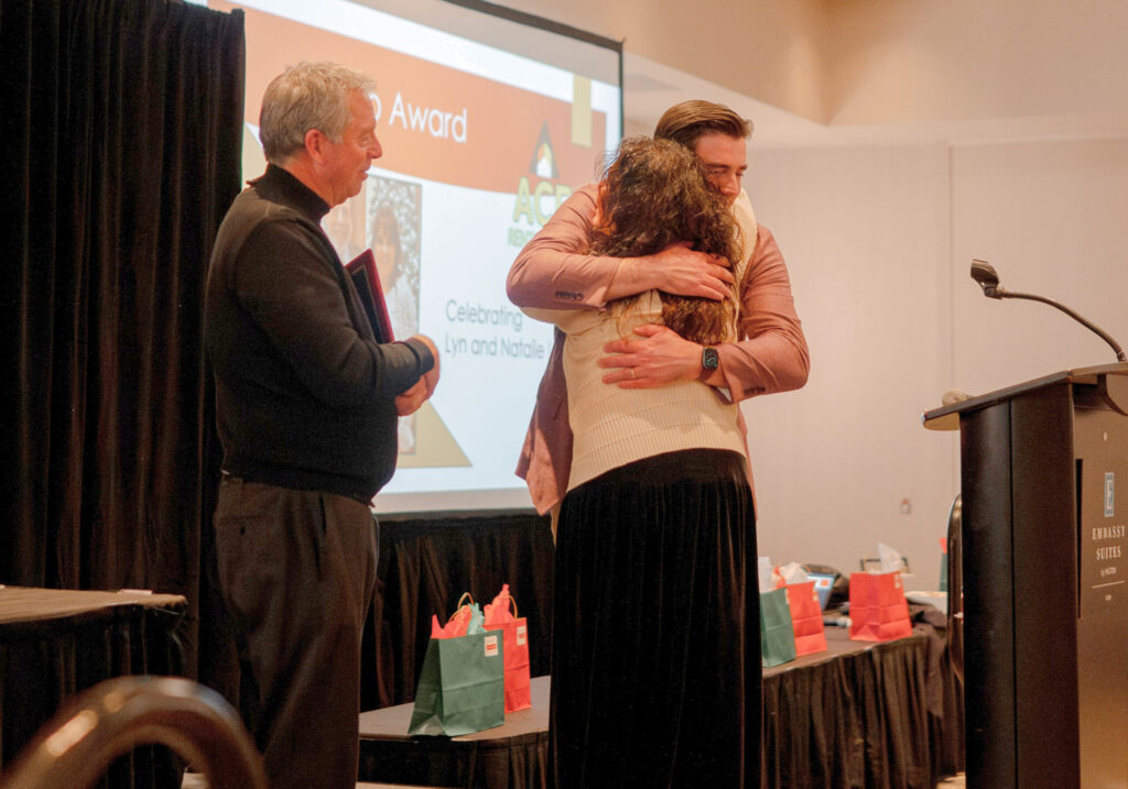 A man hugs a woman during an awards presentation while another presenter stands nearby on stage in front of a projection screen.