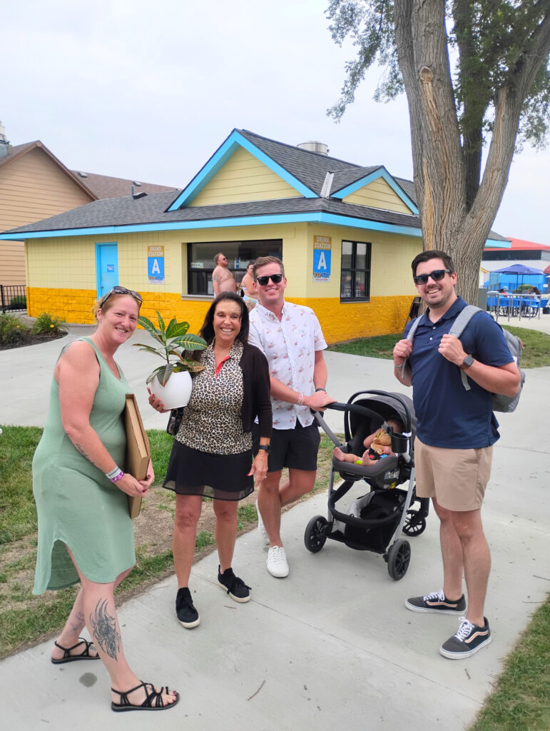 Two women two men, and a baby in a stroller pose for a photo outside with a woman holding a potted plant.
