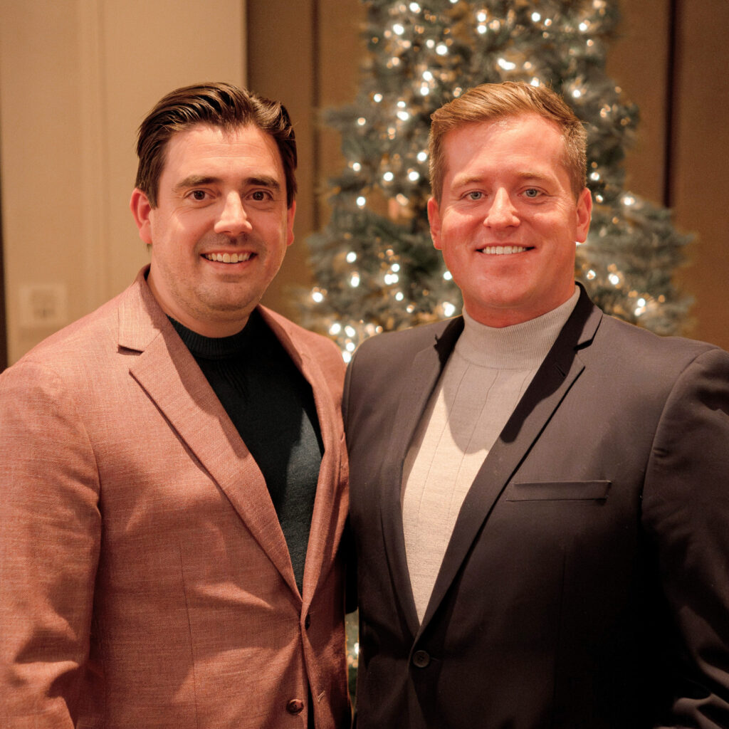 Two men in business attire smile while posing together in front of a decorated Christmas tree at an indoor event.