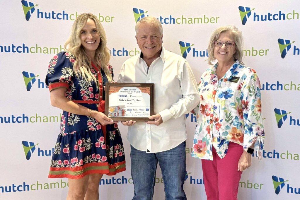 A man with white hair stands between two women – one younger, one older – holding a plaque as all smile. Behind them is a backdrop with a blue and green repeated “hutch chamber” logo all over it.