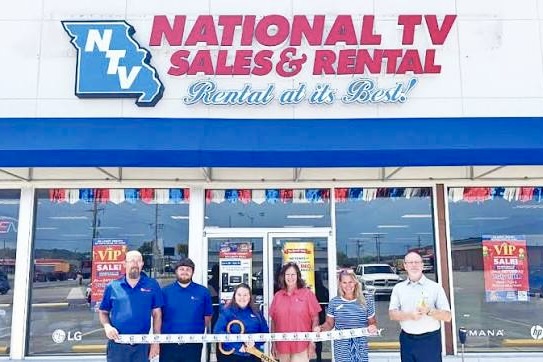 Several men and women stand lined up in front of a building with a National TV Sales & Rental sign at the top of it. They hold a long white ribbon, which is about to be cut by a woman wearing a bright blue shirt.
