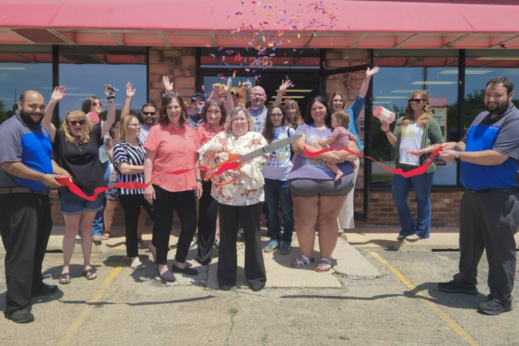 A group of men and women stand in front of a building with a long red ribbon that has just been cut by a woman holding a giant pair of scissors. Confetti falls as several people hold up their arms in excitement.