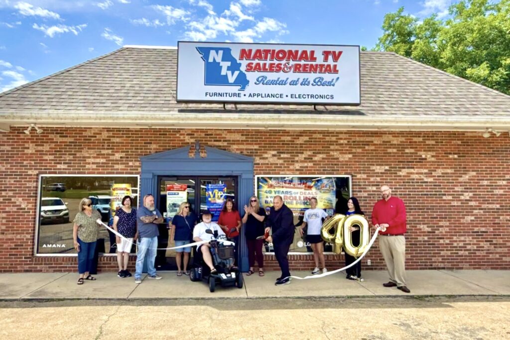 group of men and women stand lined up in front of a brick building with a National TV Sales & Rental sign at the top of it. They hold a long white ribbon, which has just been cut by a man wearing a blue shirt. One woman also holds giant gold mylar “40” balloons.