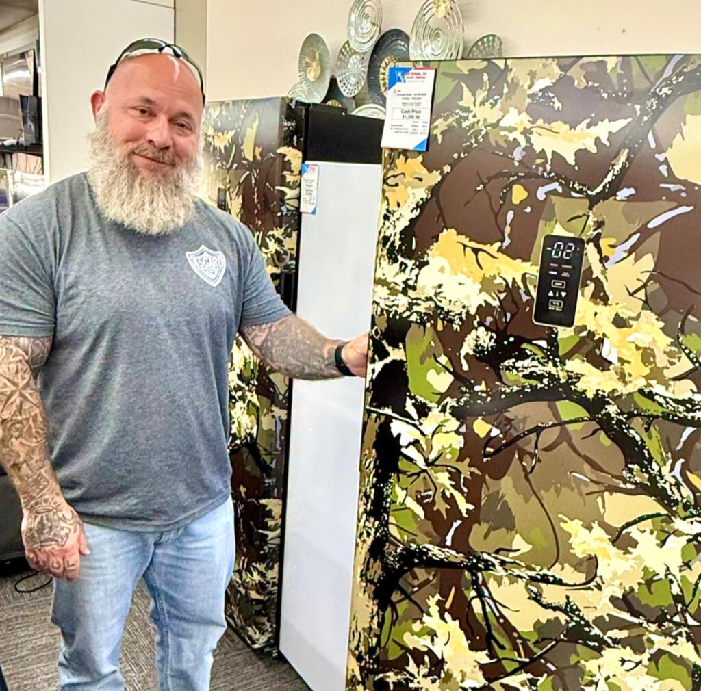 A man stands beside a refrigerator covered in army-green camouflage.