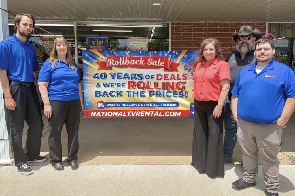 Five men and women stand on either side of a banner announcing a “rollback sale” celebrating National TV’s 40 years in business.