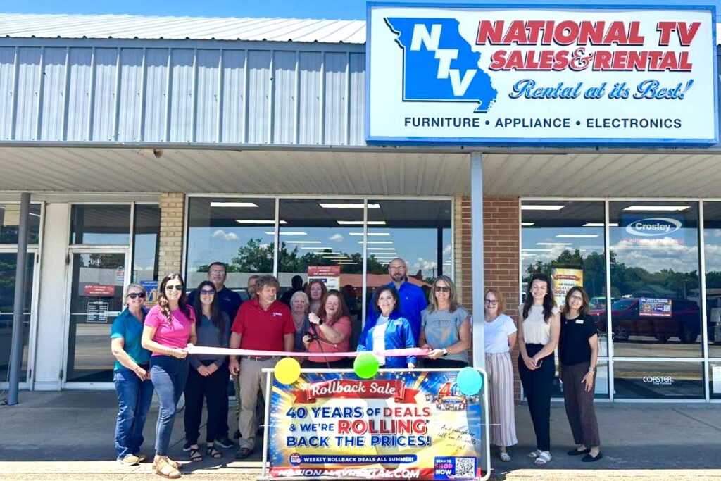 A group of men and women stand lined up in front of a brick building with a National TV Sales & Rental sign at the top of it. They hold a long pink ribbon as a woman wearing a pink shirt prepares to cut it.