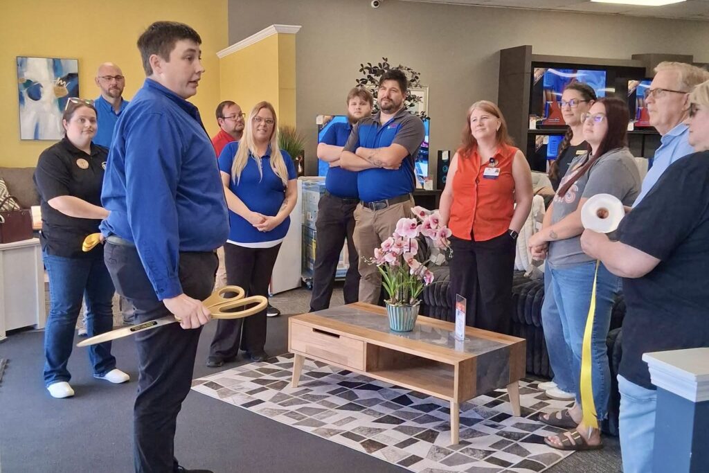 A man wearing a bright blue shirt and holding a giant pair of scissors talks to a group of men and women inside a store, one of whom holds a roll of wide yellow ribbon.