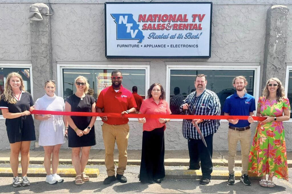 Several men and women stand lined up in front of a building with a National TV Sales & Rental sign at the top of it. They hold a long red ribbon, which is about to be cut by a man wearing a blue plaid shirt and holding a giant pair of scissors.