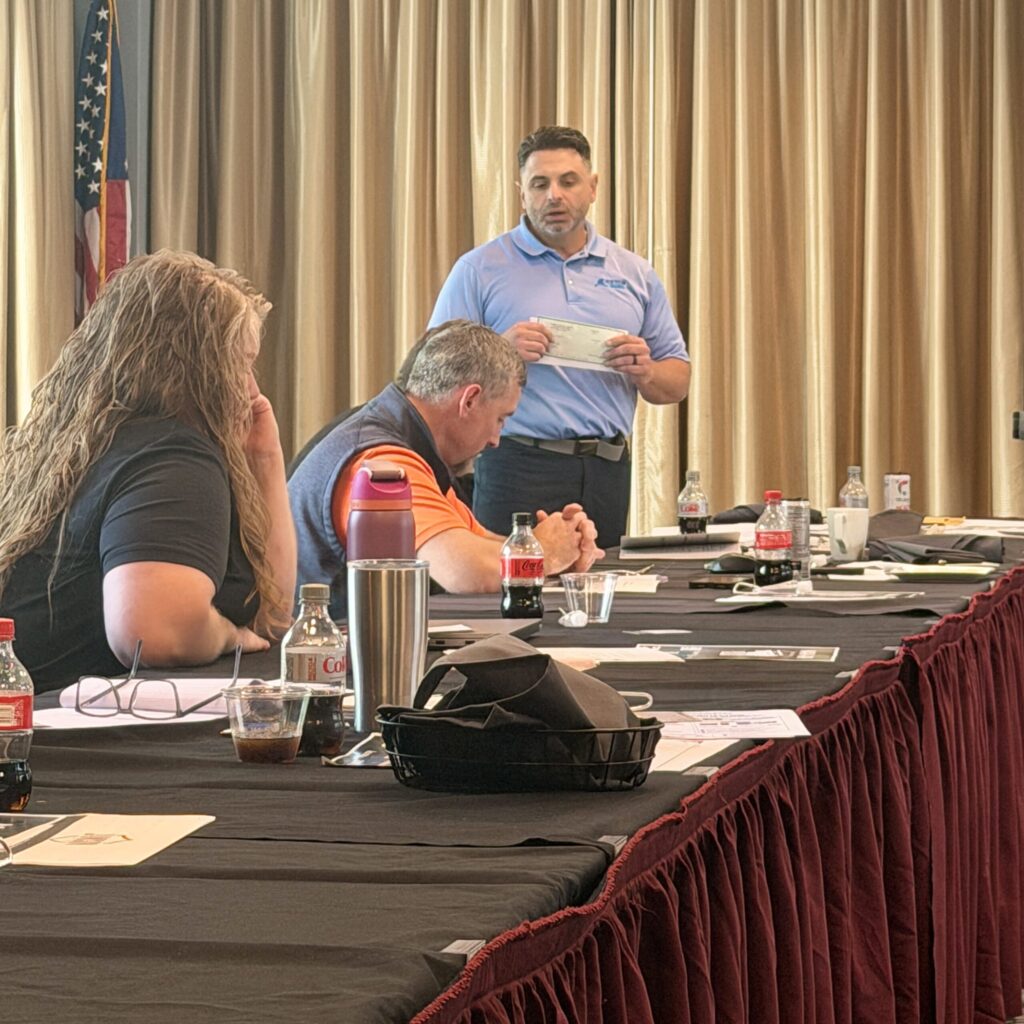 A man wearing a blue polo shirt stands at his place at a conference table, holding a check and speaking to his seated colleagues.