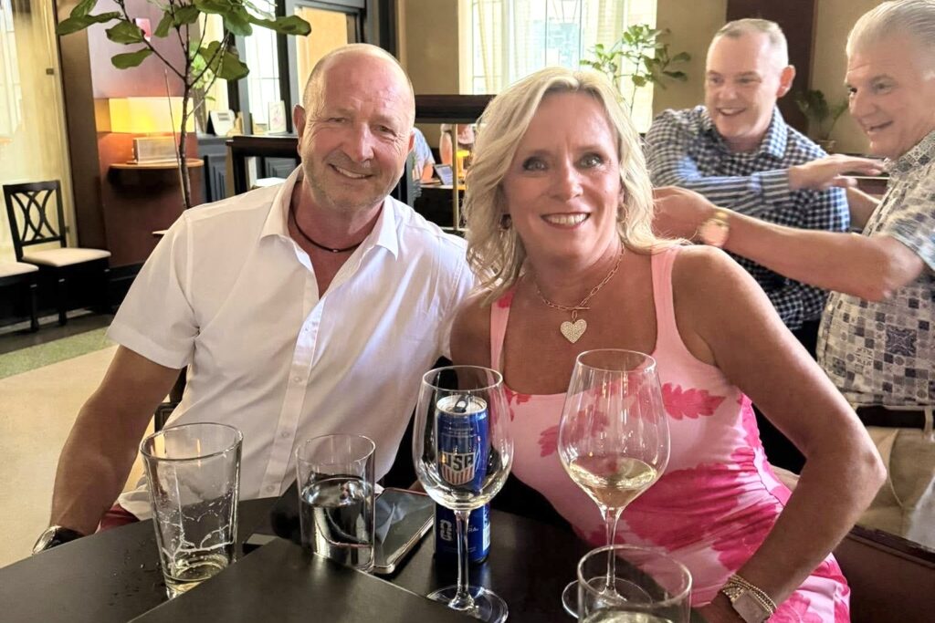 A man and a woman lean toward one another and smile broadly as they sit at a table with wineglasses. Two men behind them share a laugh.