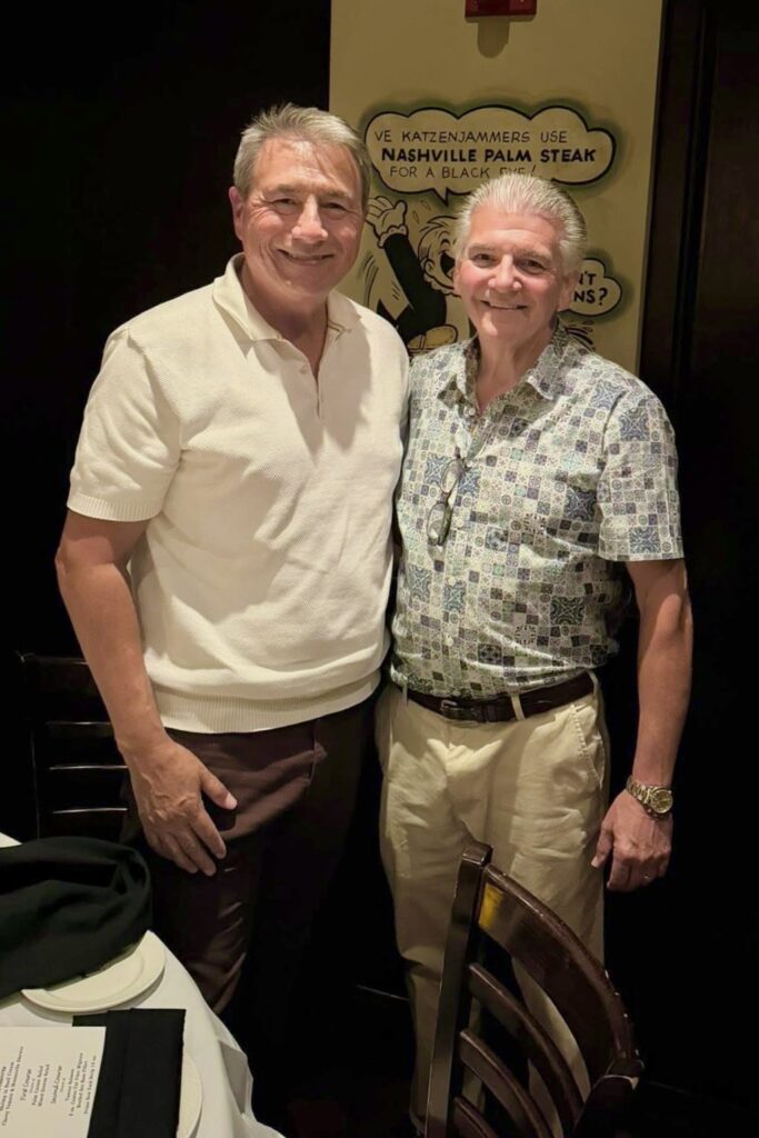 Two older men stand next to one another, smiling.