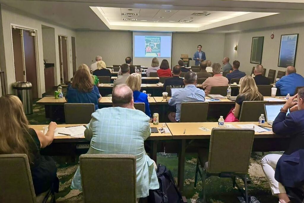 A large group of adults sit in a professional classroom setting, with a man at the front of the room giving a presentation.