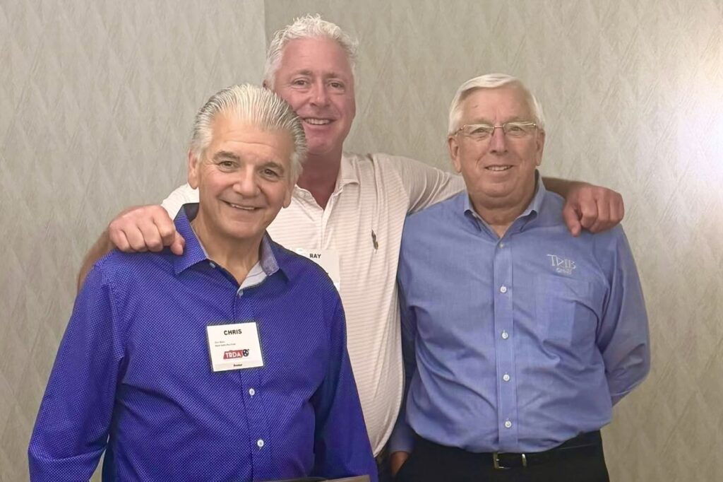 Three older men stand together, smiling.