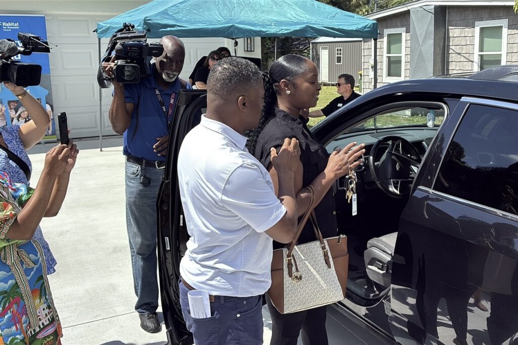 A man helps a woman with her eyes shut from a black vehicle as videographers and photographers record the event.