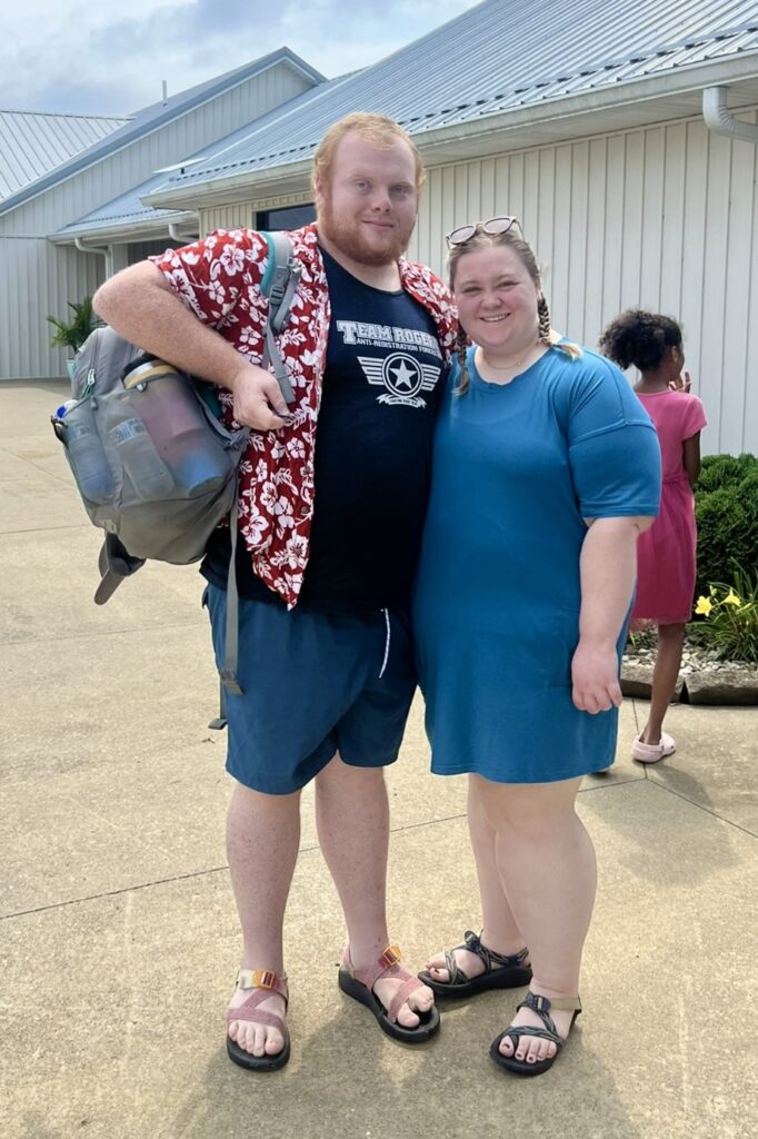 A man and woman wearing summery clothing and sandals, stand together outside, smiling. The man totes an overstuffed backpack.
