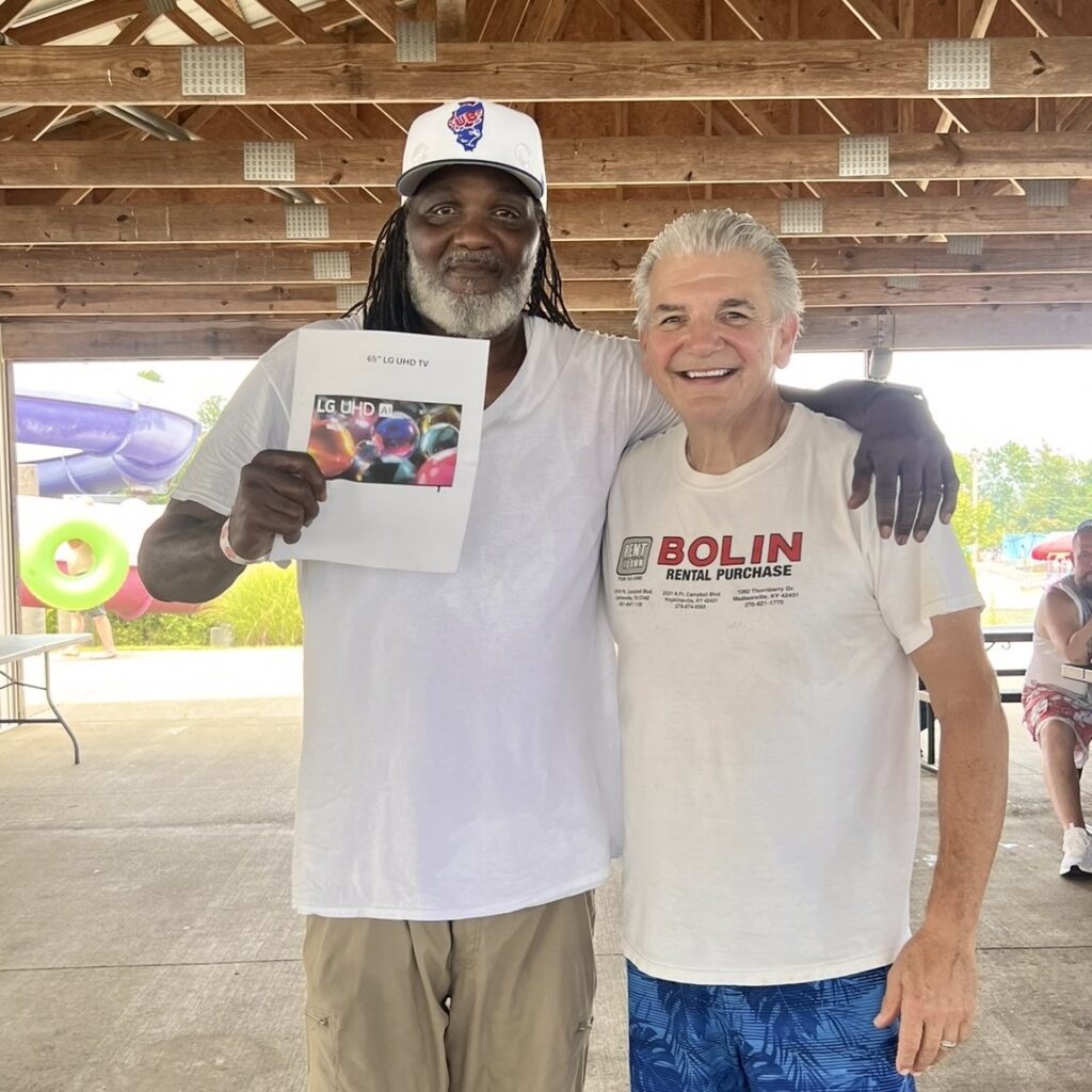 Two men stand outside together smiling. One man is wearing a tee and khakis, and holding up a piece of paper with a picture of an LG TV on it; the other is wearing a white Bolin Rental Purchase tee and blue swim trunks.