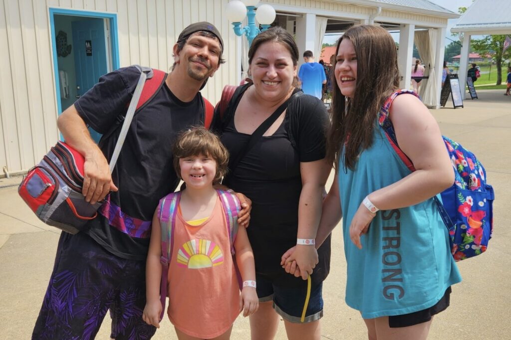 A man, woman, and two children, dressed in summer clothing and swimwear and carrying backpacks and totes, stand together outside and smile.