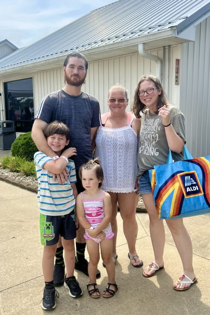 A man, woman, and three children, dressed in summer clothing and swimwear and carrying backpacks and totes, stand outside together and smile.