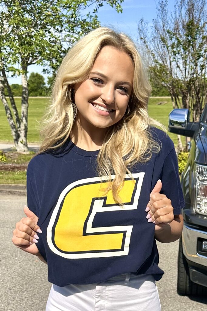 A young blonde woman wearing a navy tee with a yellow “C” logo smiles and gives a thumbs up.
