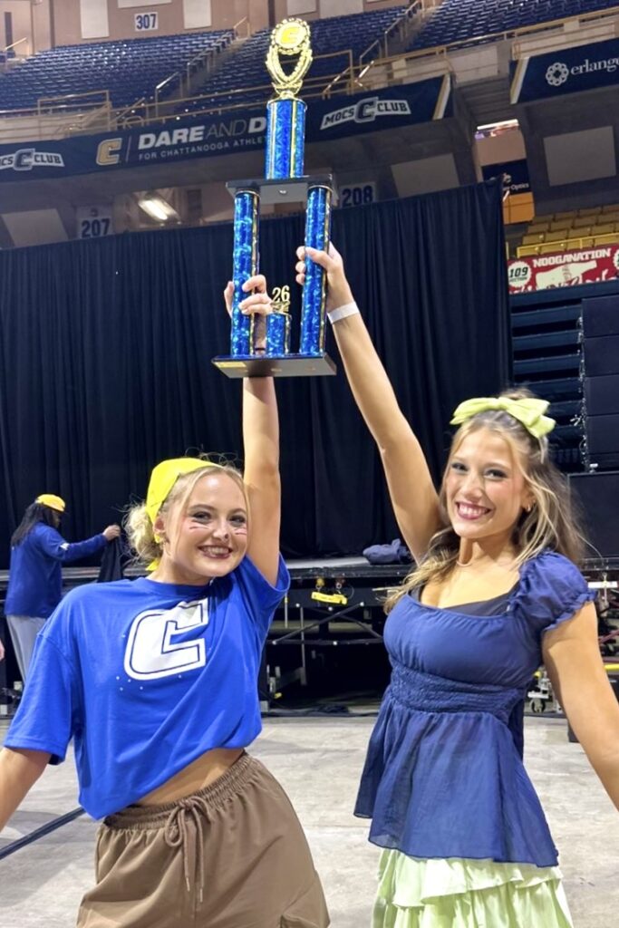 Two young women stand together in an arena, triumphantly holding up a large trophy and smiling broadly.