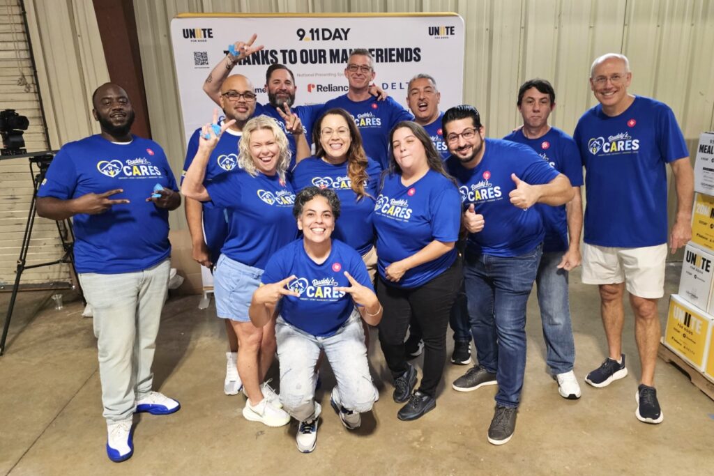 A group of people wearing matching blue Buddy’s Cares tees smile with a sign behind that reads “9/11 Day – Thanks to our Many Friends.”