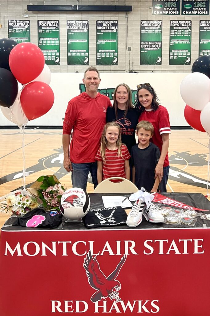 A young woman wearing a black “Montclair Volleyball” tee stands smiling in a gymnasium with her arms around a bearded man and brunette woman, behind a young boy and young girl. Before them is a table full of black and red celebratory paraphernalia, with a huge red banner reading “Montclair State Redhawks.”