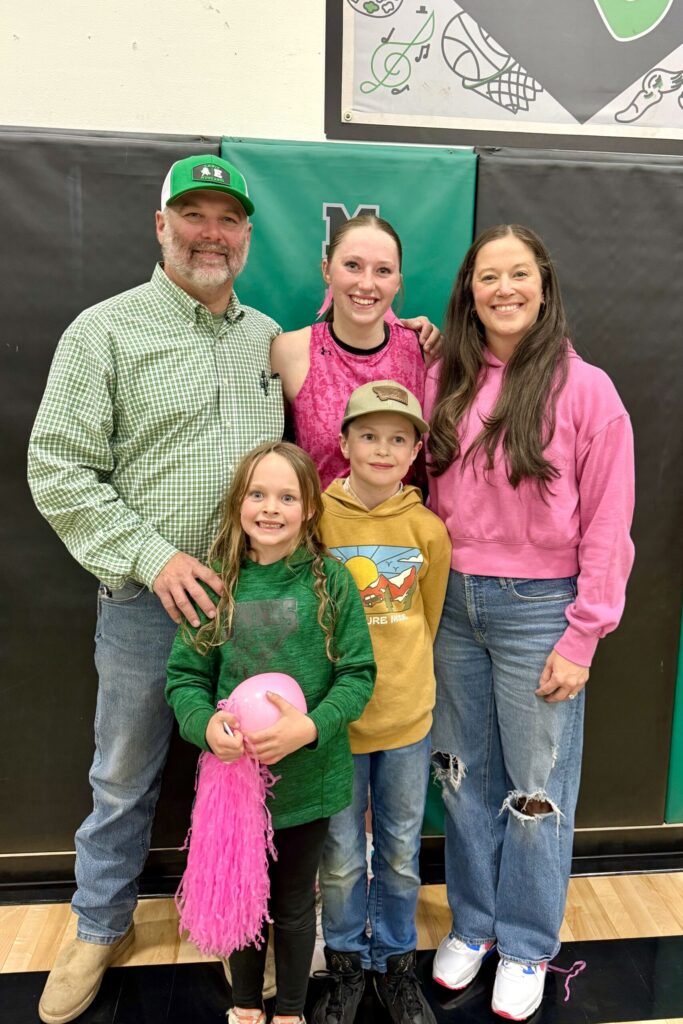 A young woman wearing a bright pink tee stands smiling in a gymnasium with her arms around a bearded man and brunette woman, behind a young boy and young girl holding a pompom and a balloon.