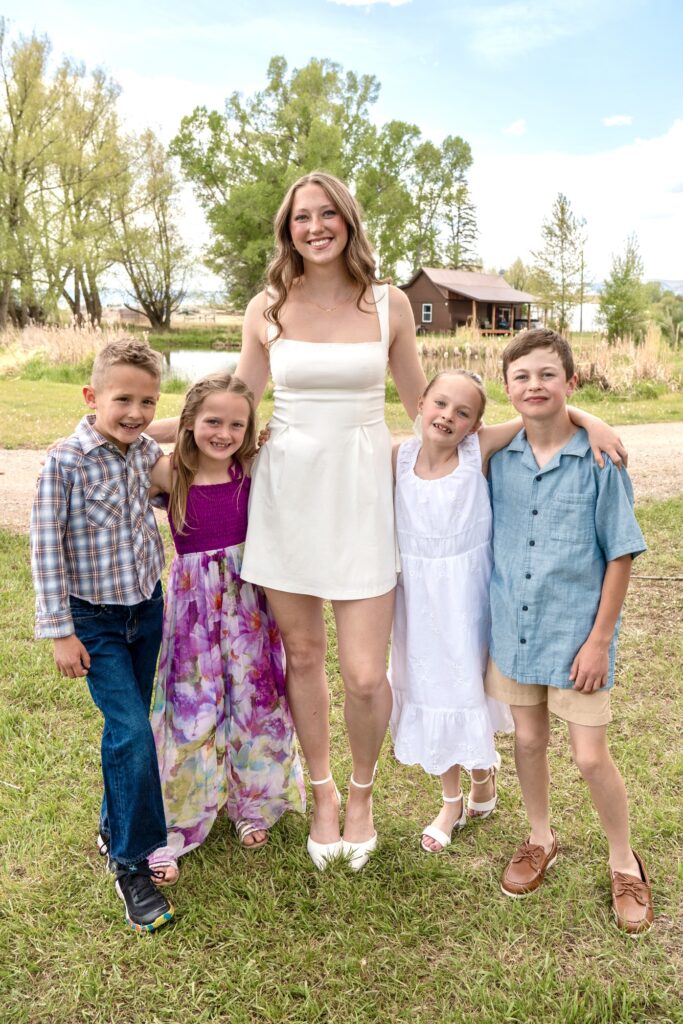 A young woman in a white sundress stands outside with two young boys and two young girls on either side of her, all well-dressed and smiling.