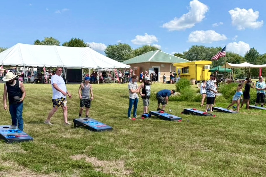A line of people beside cornhole boards participate in the game in a park environment.