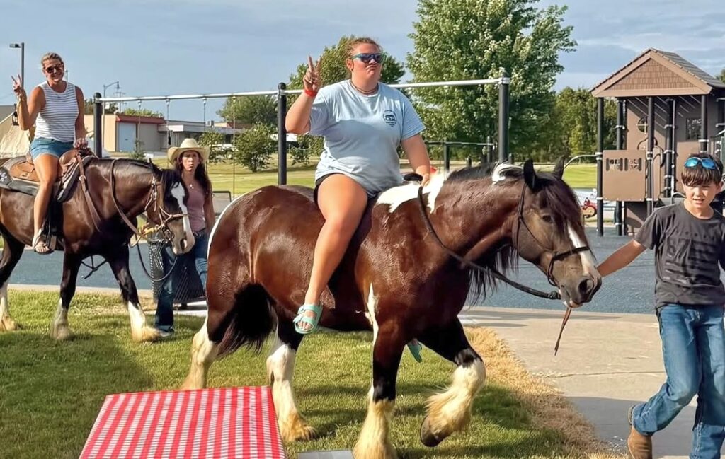 Two women ride chestnut-colored horses being led by a woman and a young boy through a park.