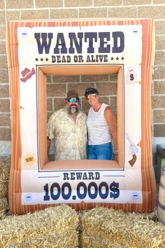 A middle-aged couple wearing sunglasses stand smiling behind a huge inflatable frame reading “Wanted Dead or Alive / Reward $100,000.”