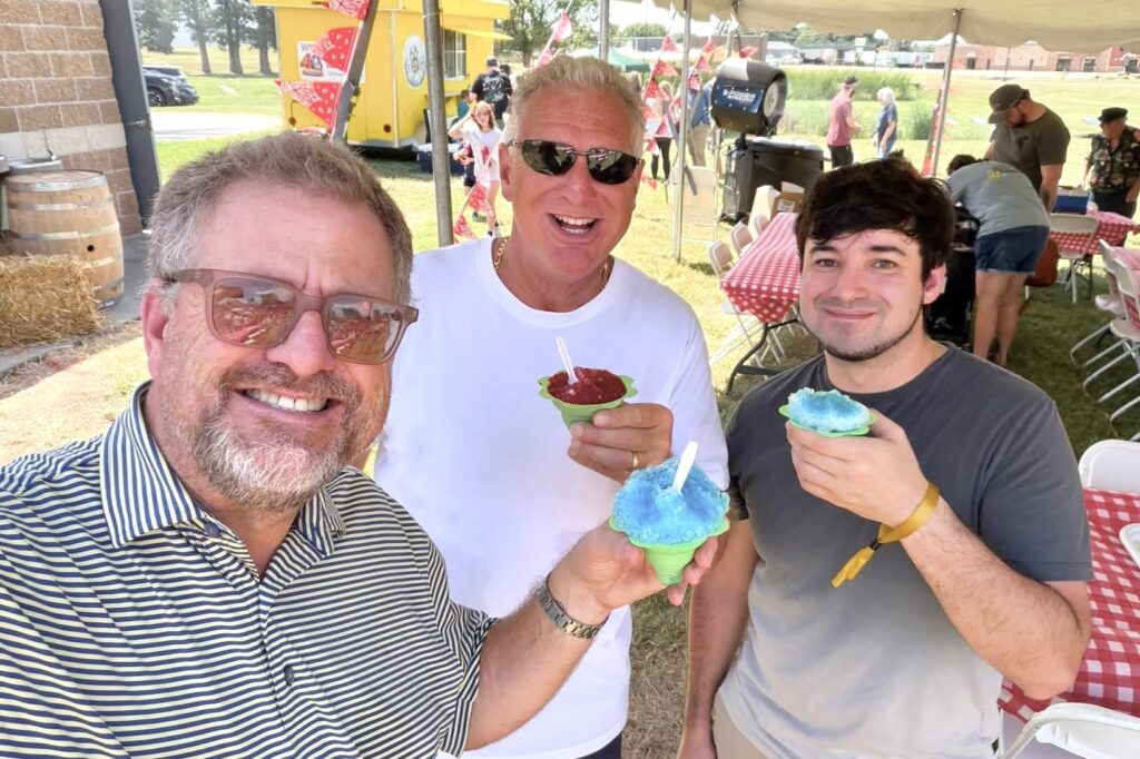 Three men standing under the shade of a white tent smile broadly while holding up colorful snow cones.
