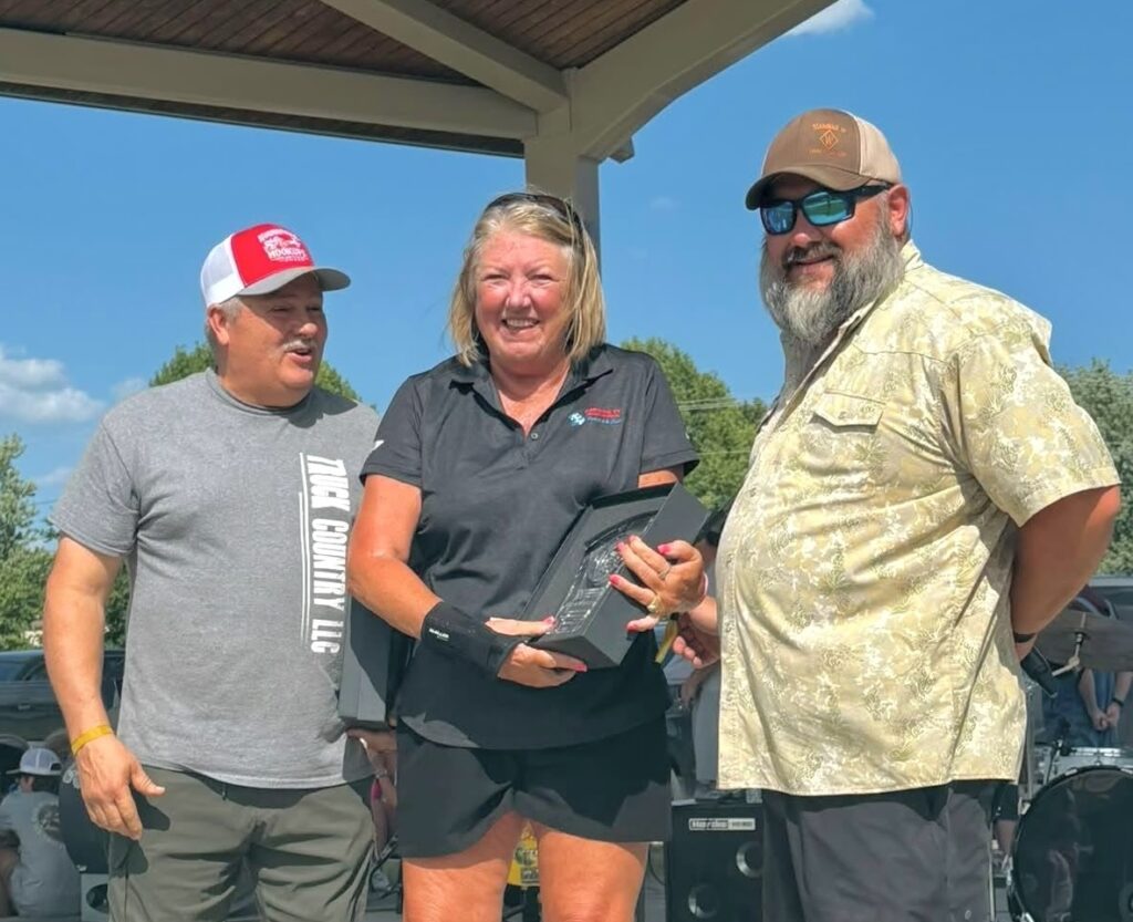 Three middle-aged people – a woman between two men – wearing summer clothing stand smiling in the sunshine. The men both wear baseball caps and sunglasses, while the woman holds a black box with a crystal clock inside it.
