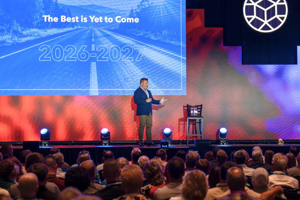 A man in chinos and a blue jacket stands up on stage presenting to a large crowd. A big screen behind him reads “The Best is Yet to Come | 2026-2027.”