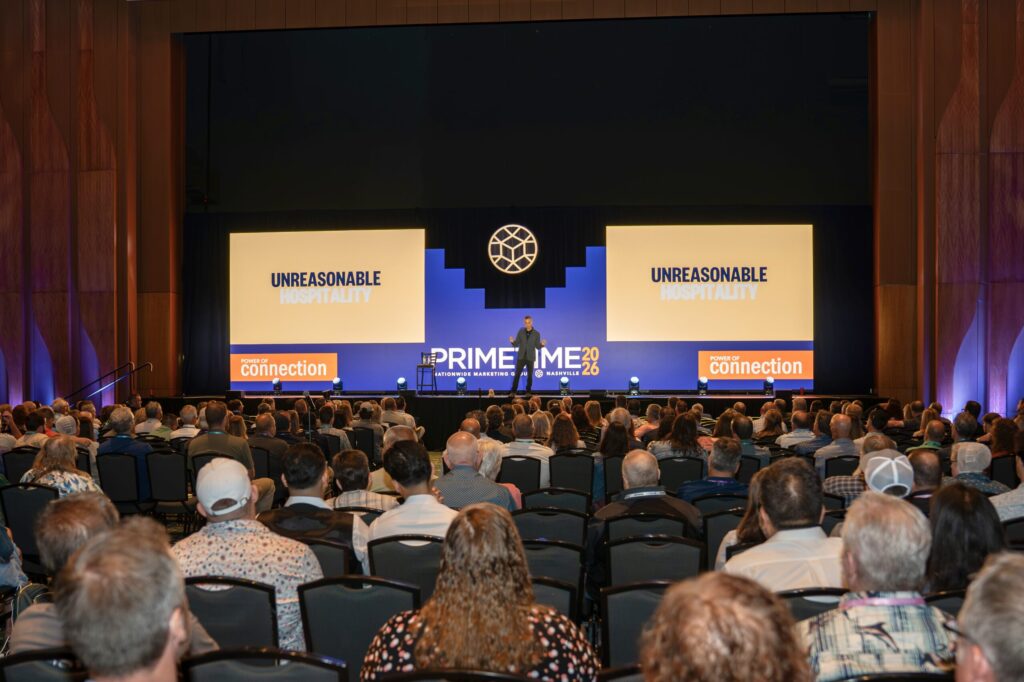 A man in dark gray pants and jacket stands up on an expansive stage presenting to a large audience. Two big screens behind him read “Unreasonable Hospitality.”