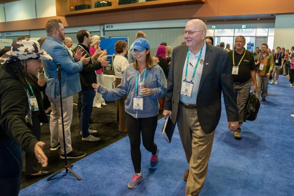 A stream of people walk along a blue carpet smiling as others applaud and high-five them from the sides.