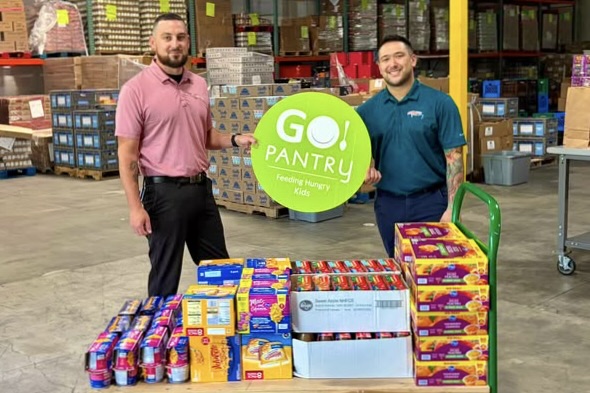 Two men smile as they hold up a lime green GO! Pantry sign, with a pallet of canned goods in front of them and a warehouse full of food behind them.