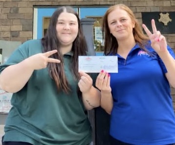 Two women stand under a Boys & Girls Clubs of Washington County sign, smiling and holding a check.