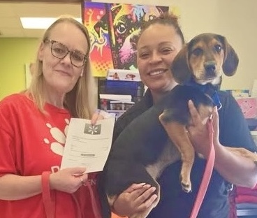 Two women stand together smiling, one holding a piece of paper and the other holding a black-and-brown-colored Coonhound puppy.