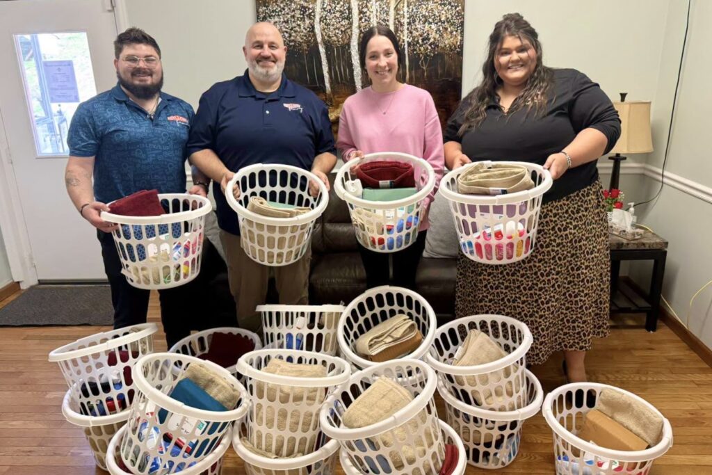 Two men and two women stand together smiling and holding small laundry baskets full of supplies, with many more baskets piled in front of them on the ground.
