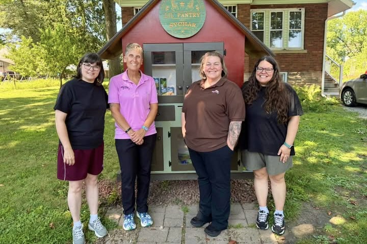 Four women stand smiling in front of a large red shed-like building with a sign reading “Lil Pantry.”