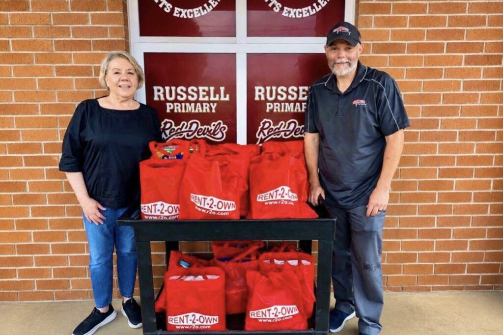 A man and woman stand on either side of a large cart holding many red bags full of school supplies. Behind them are signs reading “Russell Primary Red Devils.”